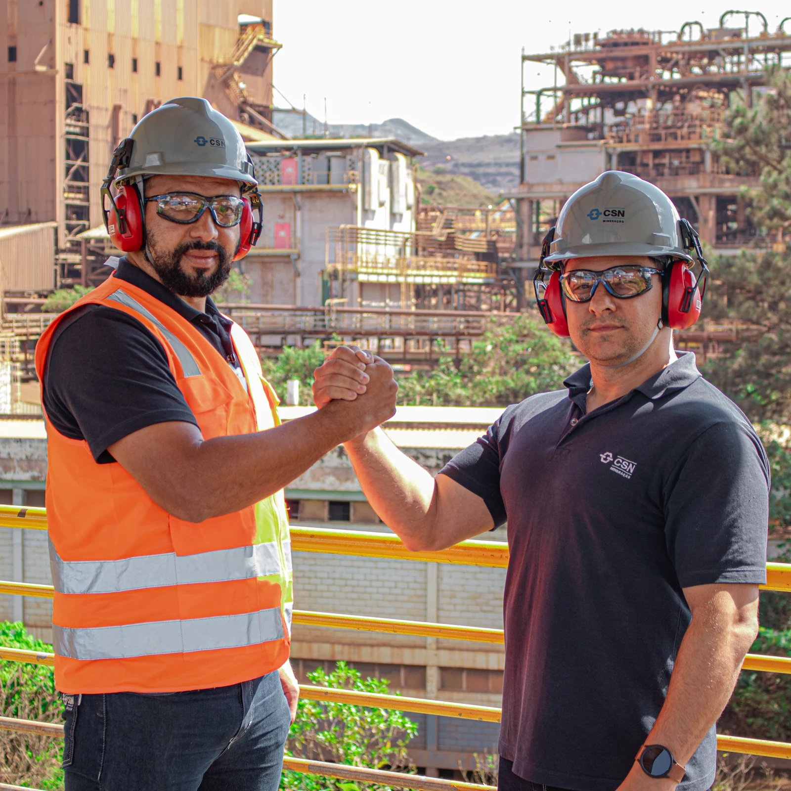 Dois colaboradores da CSN Mineração, de capacete e protetor auricular, se cumprimentando no guarda-corpo da usina